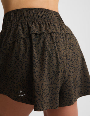 Close-up of a person wearing high-waisted athletic shorts with an allover dark leopard print on a muted olive-brown base. The shorts feature an elastic waistband, a ruffled waist detail, and a small white stylized logo near the lower left hem.