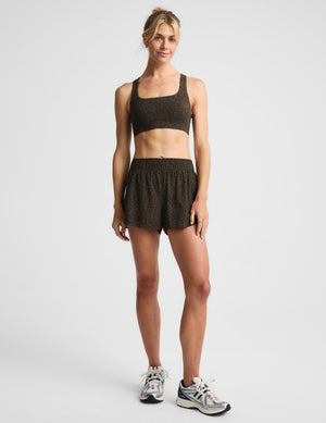 A woman modeling a matching dark brown and black speckled activewear set, consisting of a sports bra and shorts, paired with gray running shoes, against a light gray studio background.
