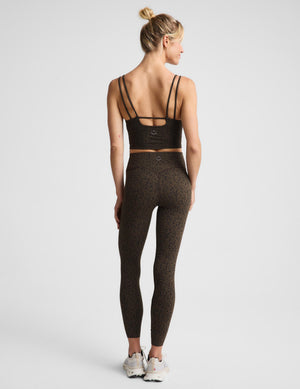 A woman posing in a matching dark brown and black speckled activewear set, featuring a sports bra and leggings against a plain light gray background.