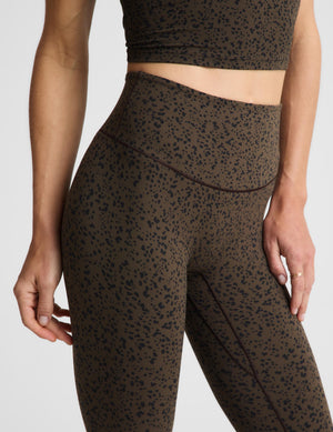 A woman posing in a matching dark brown and black speckled activewear set, featuring a sports bra and leggings against a plain light gray background.