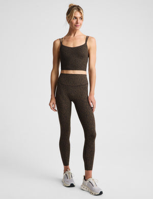 A woman posing in a matching dark brown and black speckled activewear set, featuring a sports bra and leggings against a plain light gray background.