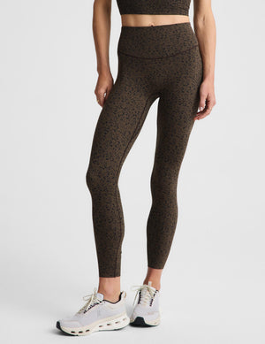 A woman posing in a matching dark brown and black speckled activewear set, featuring a sports bra and leggings against a plain light gray background.
