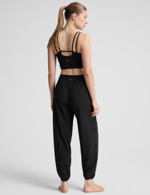 A woman modeling a black two-piece activewear set consisting of a cropped tank top and high-waisted jogger pants, photographed against a neutral light gray studio background.