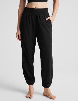 A woman modeling a black two-piece activewear set consisting of a cropped tank top and high-waisted jogger pants, photographed against a neutral light gray studio background.