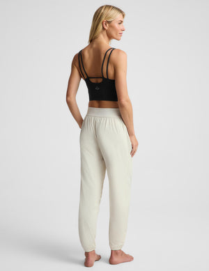 A woman stands barefoot against a light gray studio background wearing a black cropped tank top and off-white jogger-style pants with a wide elastic waistband and side pockets.
