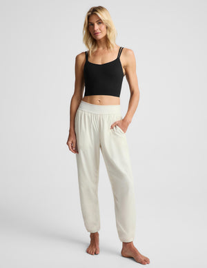 A woman stands barefoot against a light gray studio background wearing a black cropped tank top and off-white jogger-style pants with a wide elastic waistband and side pockets.