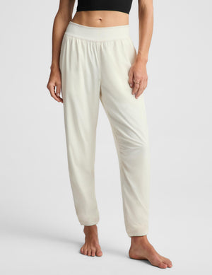 A woman stands barefoot against a light gray studio background wearing a black cropped tank top and off-white jogger-style pants with a wide elastic waistband and side pockets.