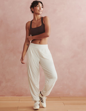 A woman modeling a pair of loose white sweatpants with a dark brown sports bra and white sneakers, standing against a textured pink wall.