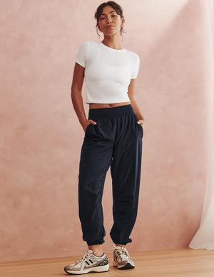 A woman stands in a studio wearing dark navy jogger-style pants with an elastic waistband and side pockets, paired with a white short-sleeve crop top and patterned sneakers, against a soft pink textured backdrop.