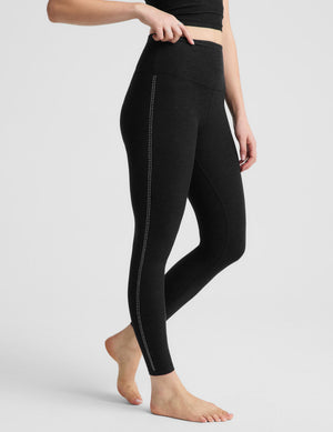A woman modeling a black athletic set consisting of a cropped tank top and high-waisted leggings, standing against a light gray studio background.