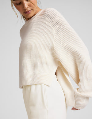 Woman wearing a beige hi lo sweater against a light gray background. 