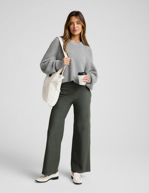 Woman wearing a gray sweater and green pants, holding a white bag and a coffee cup on a light gray background. 