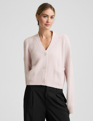 Woman wearing a light pink button-up cardigan and black pants against a plain background. 