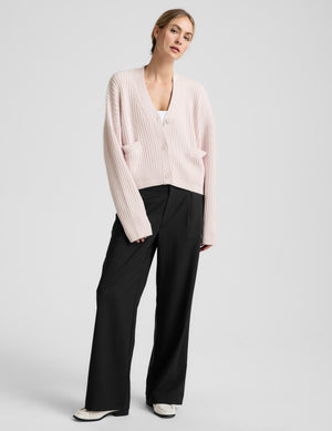 Woman wearing a light pink button-up cardigan and black pants against a plain background. 