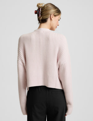 Woman wearing a light pink button-up cardigan and black pants against a plain background. 