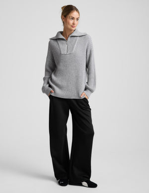 Woman wearing a gray ribbed quarter zip sweater and black pants on a light gray background. 