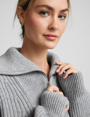 Woman wearing a gray ribbed quarter zip sweater and black pants on a light gray background. 