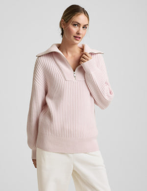 Woman wearing a light pink sweater with a zipper on a plain background. 