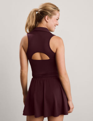 A woman shown from the back wearing a sleeveless dark burgundy athletic top with a high collar and a cutout detail at the back, set against a plain light gray studio background.