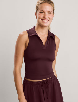 A smiling blonde woman wearing a sleeveless collared knit top in deep burgundy, paired with matching drawstring skirt, against a plain light gray studio background.