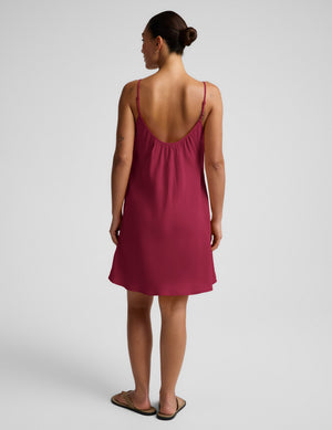 Woman wearing a magenta cupro mini dress and brown sandals against a plain background. 