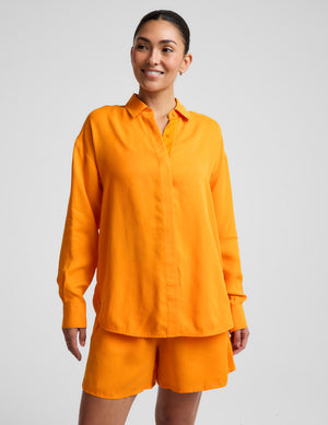 Woman wearing an orange button down shirt and orange shorts against a plain background. 