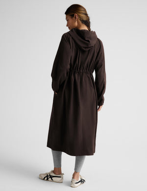 A woman modeling a dark brown hooded longline coat with drawstrings and large front patch pockets, paired with light gray leggings and white sneakers, against a light gray studio background.