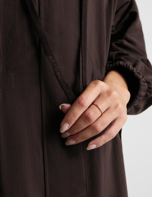 A woman modeling a dark brown hooded longline coat with drawstrings and large front patch pockets, paired with light gray leggings and white sneakers, against a light gray studio background.