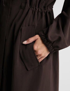 A woman modeling a dark brown hooded longline coat with drawstrings and large front patch pockets, paired with light gray leggings and white sneakers, against a light gray studio background.