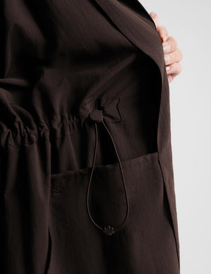A woman modeling a dark brown hooded longline coat with drawstrings and large front patch pockets, paired with light gray leggings and white sneakers, against a light gray studio background.