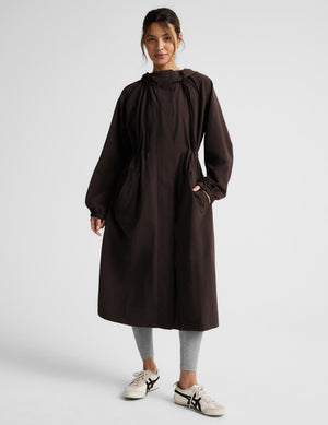 A woman modeling a dark brown hooded longline coat with drawstrings and large front patch pockets, paired with light gray leggings and white sneakers, against a light gray studio background.