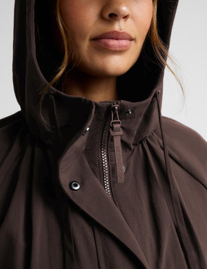 A woman modeling a dark brown hooded longline coat with drawstrings and large front patch pockets, paired with light gray leggings and white sneakers, against a light gray studio background.