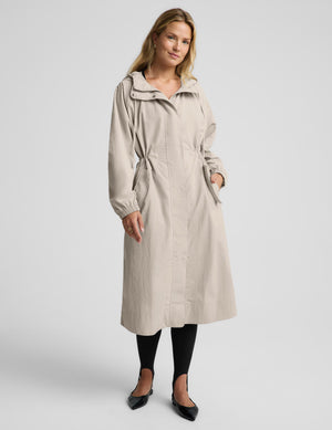 A woman modeling a light beige hooded trench coat with a tie waist, paired with black tights and black flats, against a plain light gray studio background.