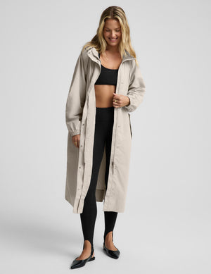 A woman modeling a long, light beige hooded raincoat over a black crop top, black leggings, and black flats, photographed against a solid light gray studio background.
