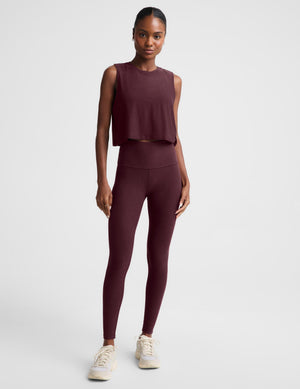 A woman modeling a sleeveless cropped tank top and matching leggings in a deep burgundy color, set against a plain light gray studio background.