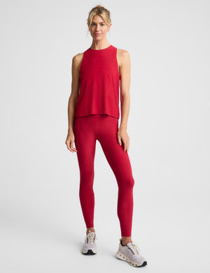 A woman modeling a matching red athletic set consisting of a sleeveless cropped tank top and high-waisted leggings, paired with light gray running shoes, against a light gray studio background.