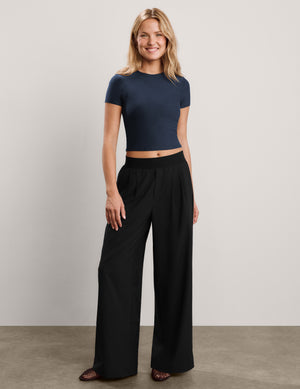A woman wearing a navy blue short-sleeve crop top and black pants, posing against a light gray studio background.