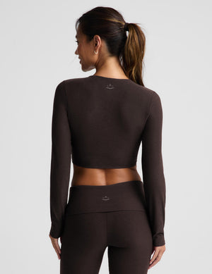 A woman wearing a cropped long sleeve top and brown foldover bootcut pants against a plain background. 
