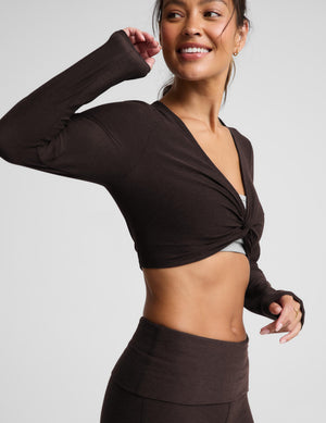 A woman wearing a cropped long sleeve top and brown foldover bootcut pants against a plain background. 