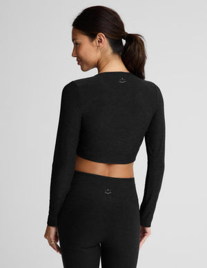 A woman modeling a black long-sleeve crop top with a twisted front detail and matching black leggings against a light gray studio background.