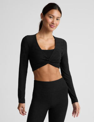 A woman modeling a black long-sleeve crop top with a twisted front detail and matching black leggings against a light gray studio background.