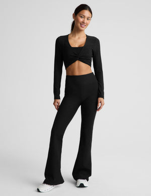 A woman modeling a black long-sleeve crop top with a twisted front detail and matching black leggings against a light gray studio background.