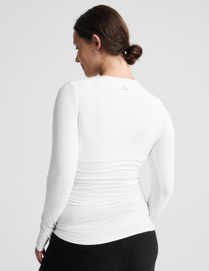A pregnant woman wearing a white long-sleeve maternity top and black leggings, standing against a light gray studio background.