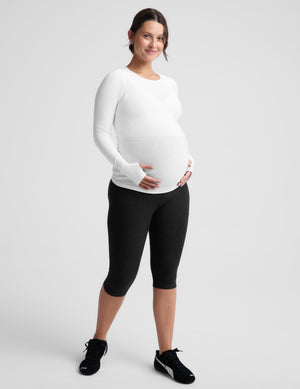 A pregnant woman wearing a white long-sleeve maternity top and black leggings, standing against a light gray studio background.