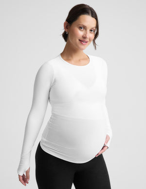 A pregnant woman wearing a white long-sleeve maternity top and black leggings, standing against a light gray studio background.