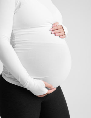 A pregnant woman wearing a white long-sleeve maternity top and black leggings, standing against a light gray studio background.