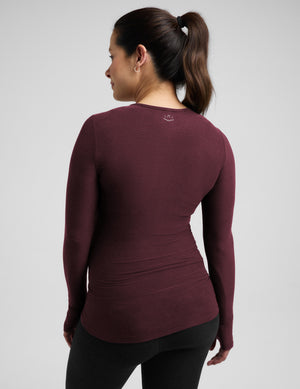 A woman shown from the back wearing a fitted long-sleeve athletic top in a deep burgundy/maroon color, paired with black leggings, against a light gray studio background.