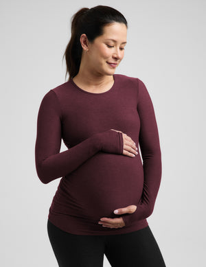 A pregnant woman wearing a long-sleeve crewneck maternity top in maroon, holding her baby bump against a solid light gray background.