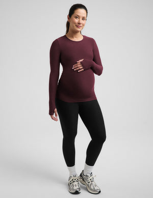 A woman modeling a long-sleeve crewneck maternity top in burgundy, paired with black leggings, white socks, and gray running shoes, against a light gray studio background.