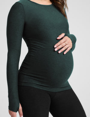 A pregnant model wearing a dark forest green long-sleeve maternity top and black leggings, holding her baby bump against a light gray background.